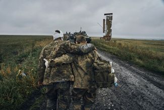 Украинский военный ведет своего раненого товарища по одной из дорог Харьковской области, недавно освобожденной украинской армией. Видна украинская военная техника и советская стела с изображением колоса и неразличимой надписью