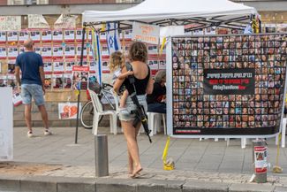 Мать, с дочерью на руках и винтовкой на плече, проходит мимо плаката с призывом вернуть заложников домой. Тель-Авив, 27 октября 2023 года