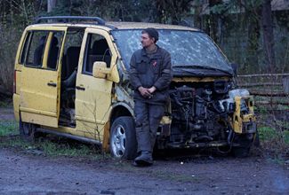 Сотрудник донецкой автомастерской стоит рядом с разбитым автомобилем. Во время обстрела погибли его коллеги